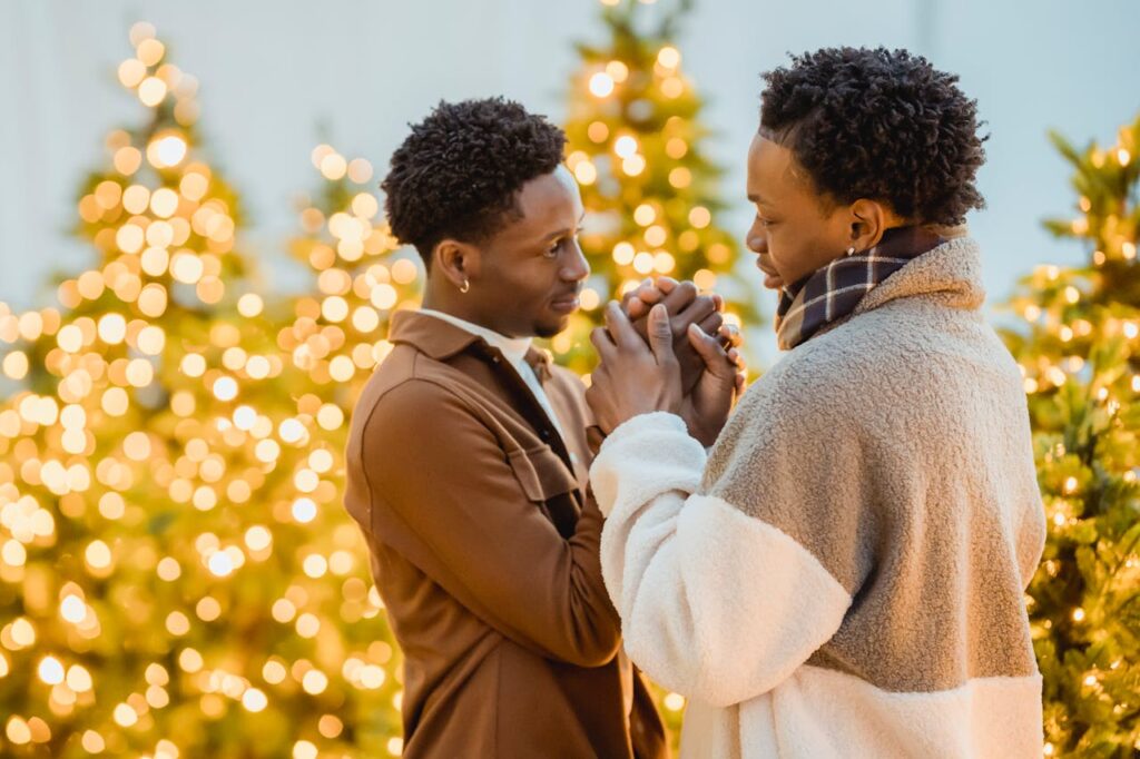 Side view of romantic African American homosexual couple holding hands and looking at each other while standing near glowing garlands on blurred background